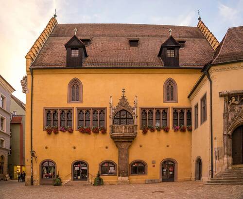 Das historische Gebäude in Regensburg zeigt kunstvolle Fenster mit Blumen, umgeben von der charmanten Altstadt und historischen Architektur.
