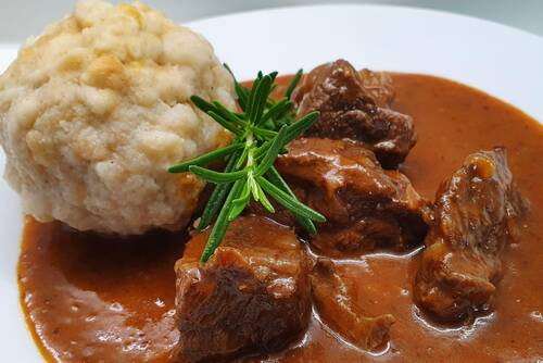 Ein Teller mit Rindergulasch, serviert mit einem Knödel und frischen Kräutern, ideal für ein gemütliches Abendessen.