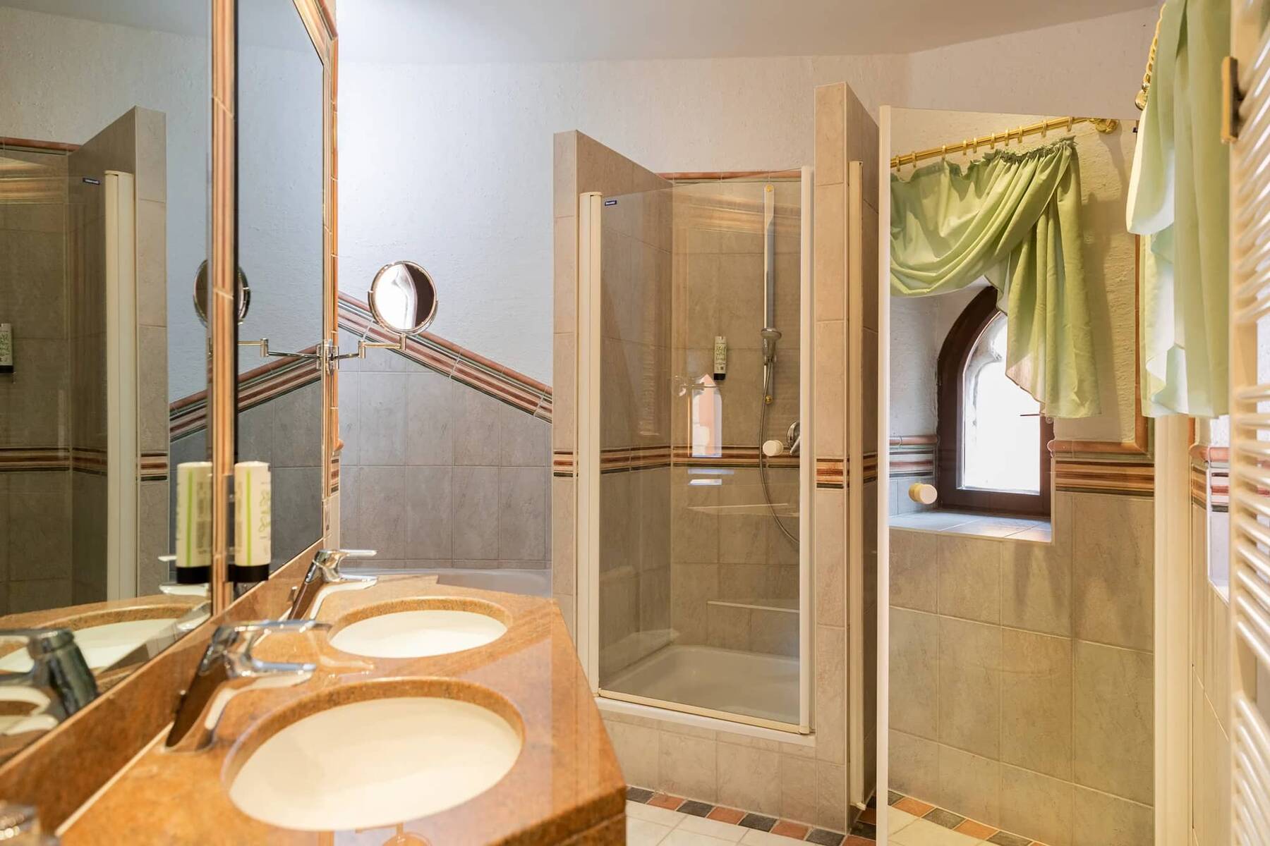 Das Badezimmer im Hotel Alpenglück zeigt eine elegante Dusche, Doppelwaschbecken und ein Fenster mit grünen Vorhängen.