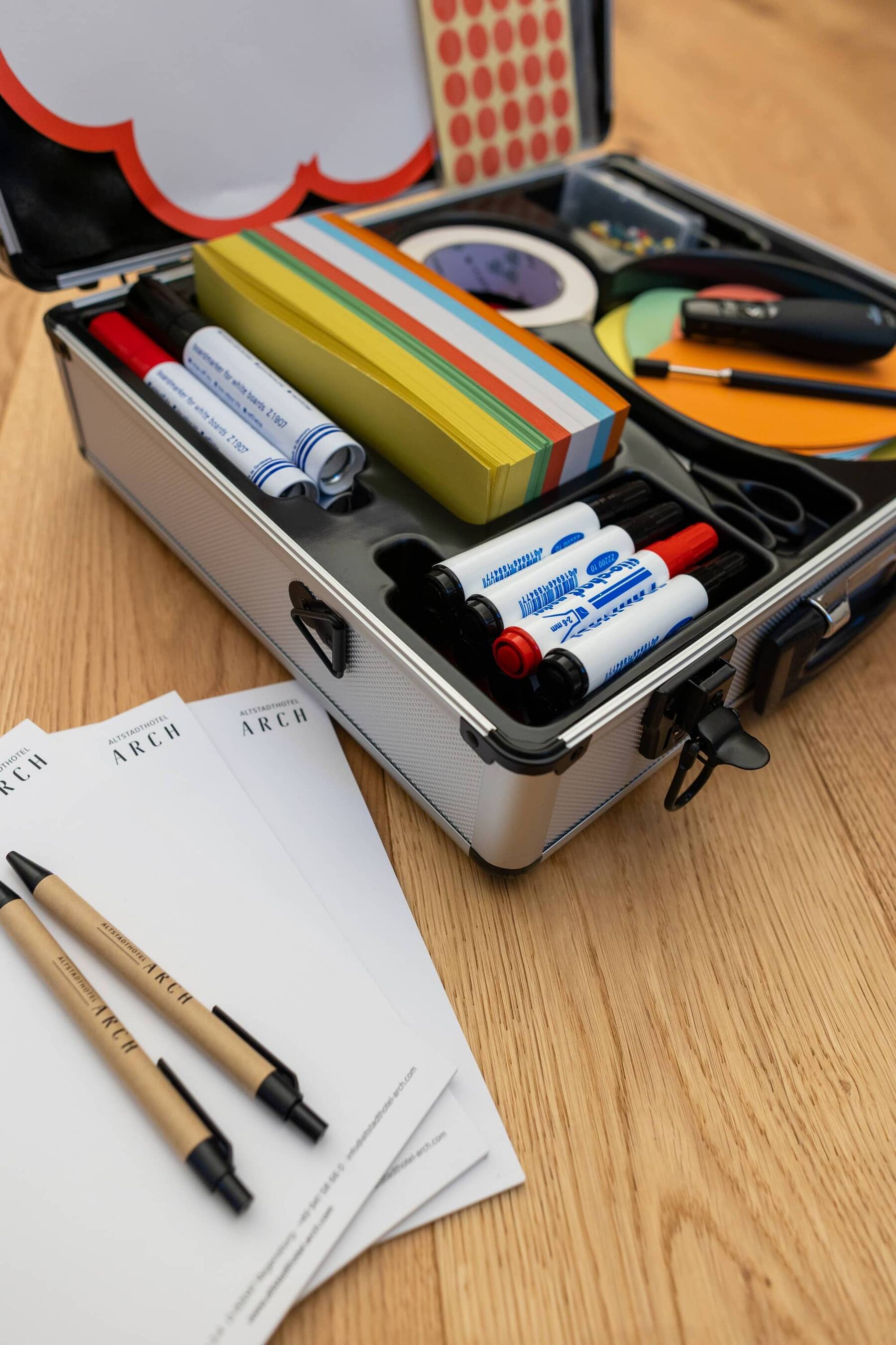 Materialkoffer mit verschiedenen Schreibutensilien, Haftnotizen und Notizblättern auf einem Holztisch für kreative Büroarbeit.