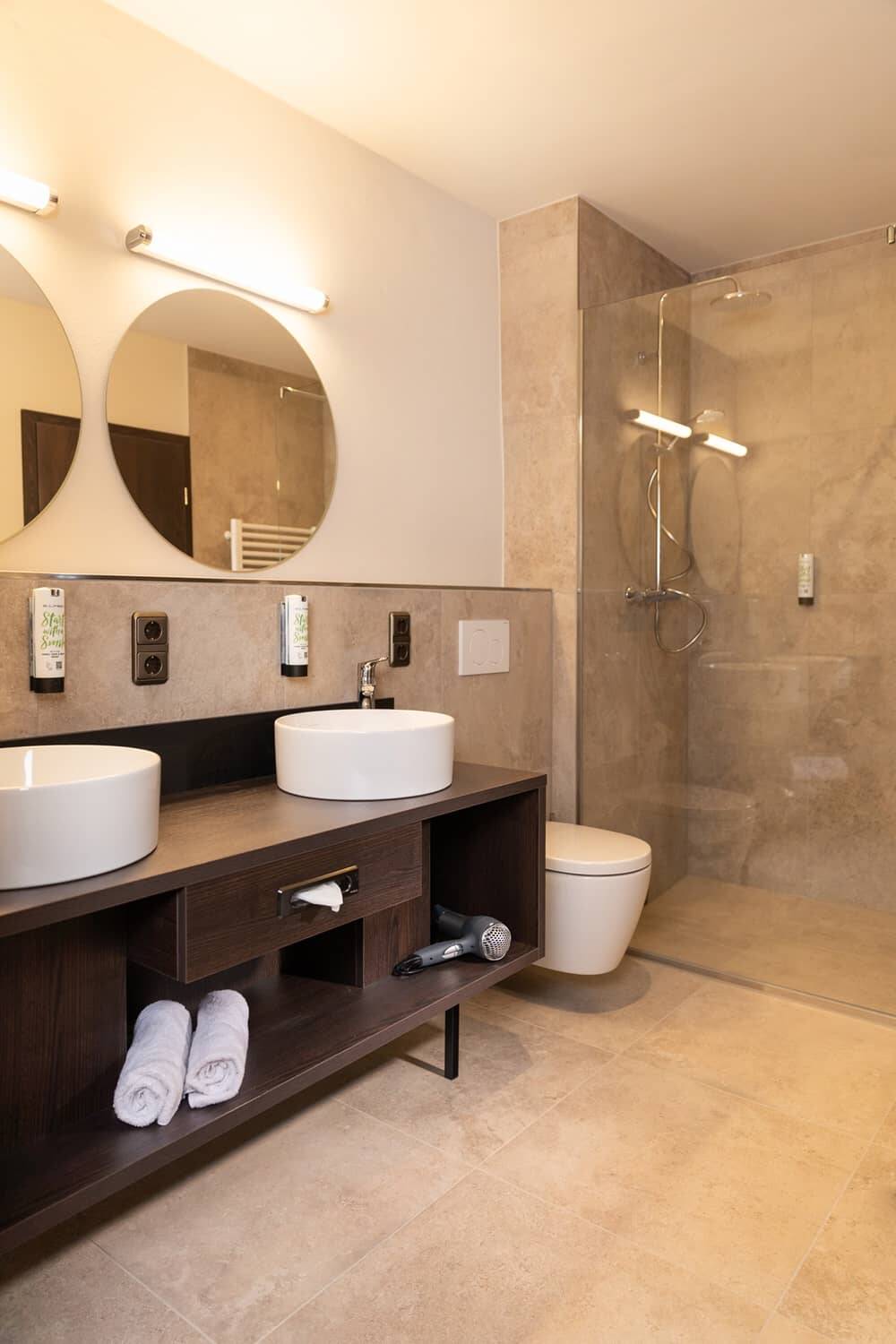 Ein modernes Badezimmer im Hotel mit zwei Waschbecken, einer Dusche und Handtüchern, ideal für einen entspannten Familienurlaub.