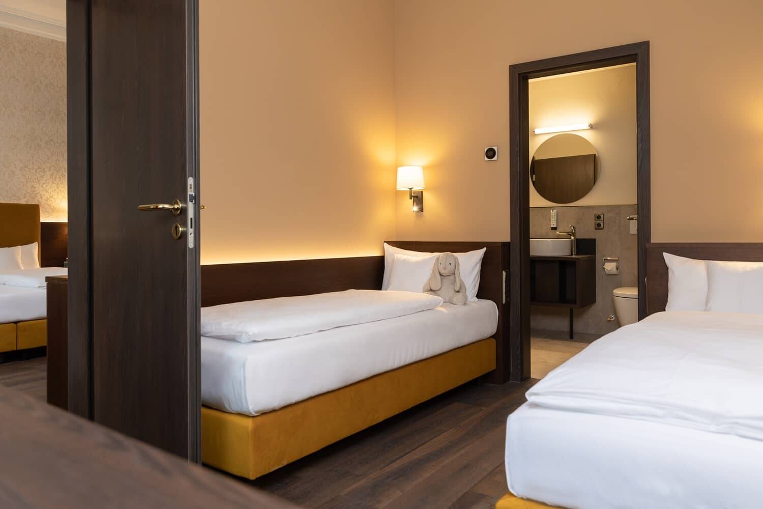 Familienzimmer im Hotel Alpenblick mit zwei Betten, einem Kuscheltier und einem Blick auf das Badezimmer. Ideal für Familienurlaube.