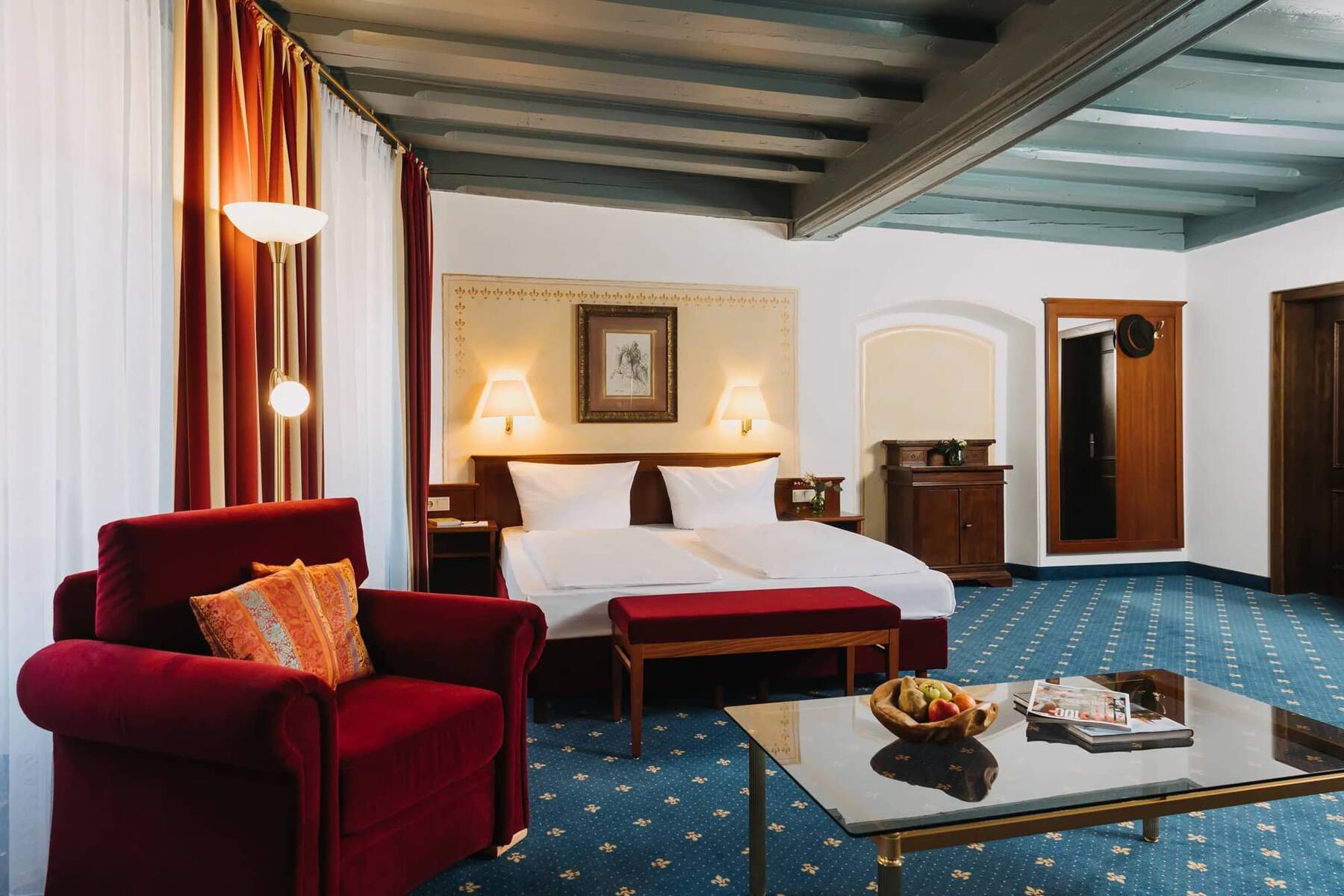 Gemütliches Zimmer im Hotel Alpenglück mit Doppelbett, rotem Sessel und Tisch, ideal für einen entspannten Familienurlaub.