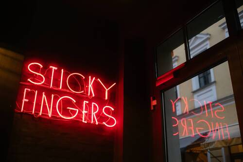 Das neonfarbene Schild „Sticky Fingers“ leuchtet in einem stilvollen Restaurant und spiegelt sich in einem Fenster.