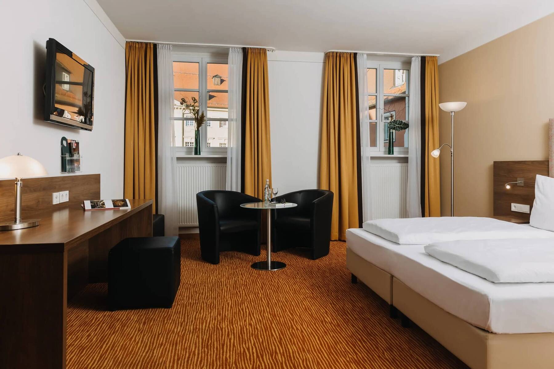Ein helles Hotelzimmer mit einem Doppelbett, einem Tisch, zwei Stühlen und großen Fenstern, ideal für einen Familienurlaub.