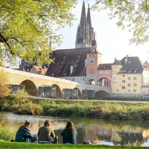 Drei Personen sitzen am Ufer des Donauufers in Regensburg, mit Blick auf die historische Altstadt und die Brücke.