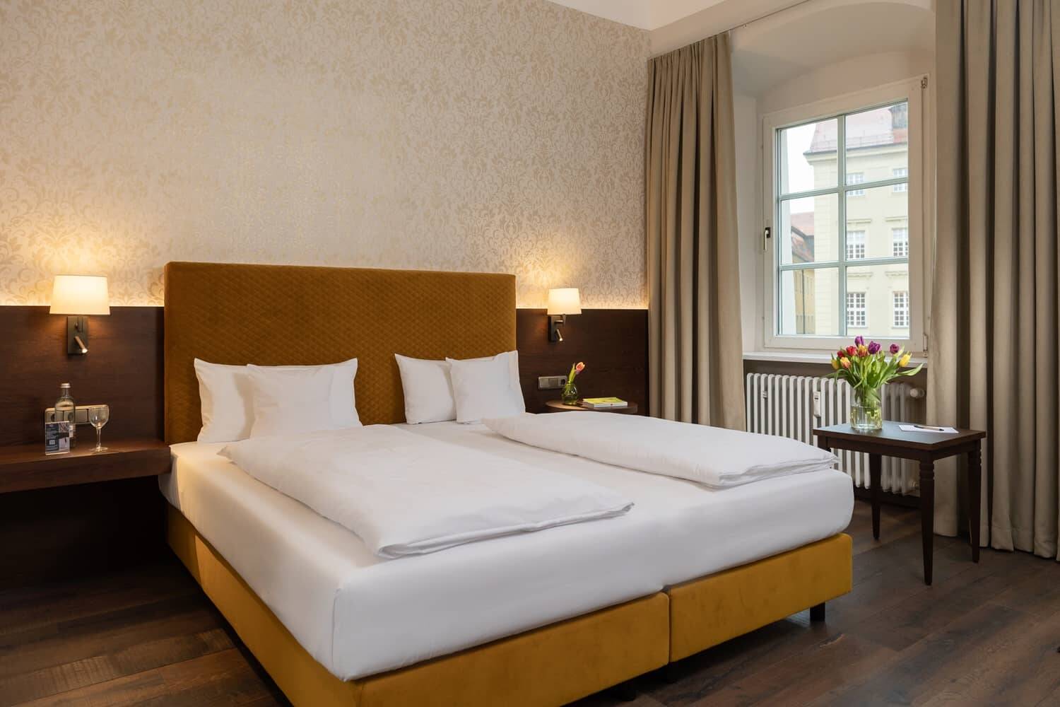 Doppelzimmer im Hotel mit einem großen Bett, stilvollen Möbeln und einem Fenster, das Tageslicht hereinlässt.