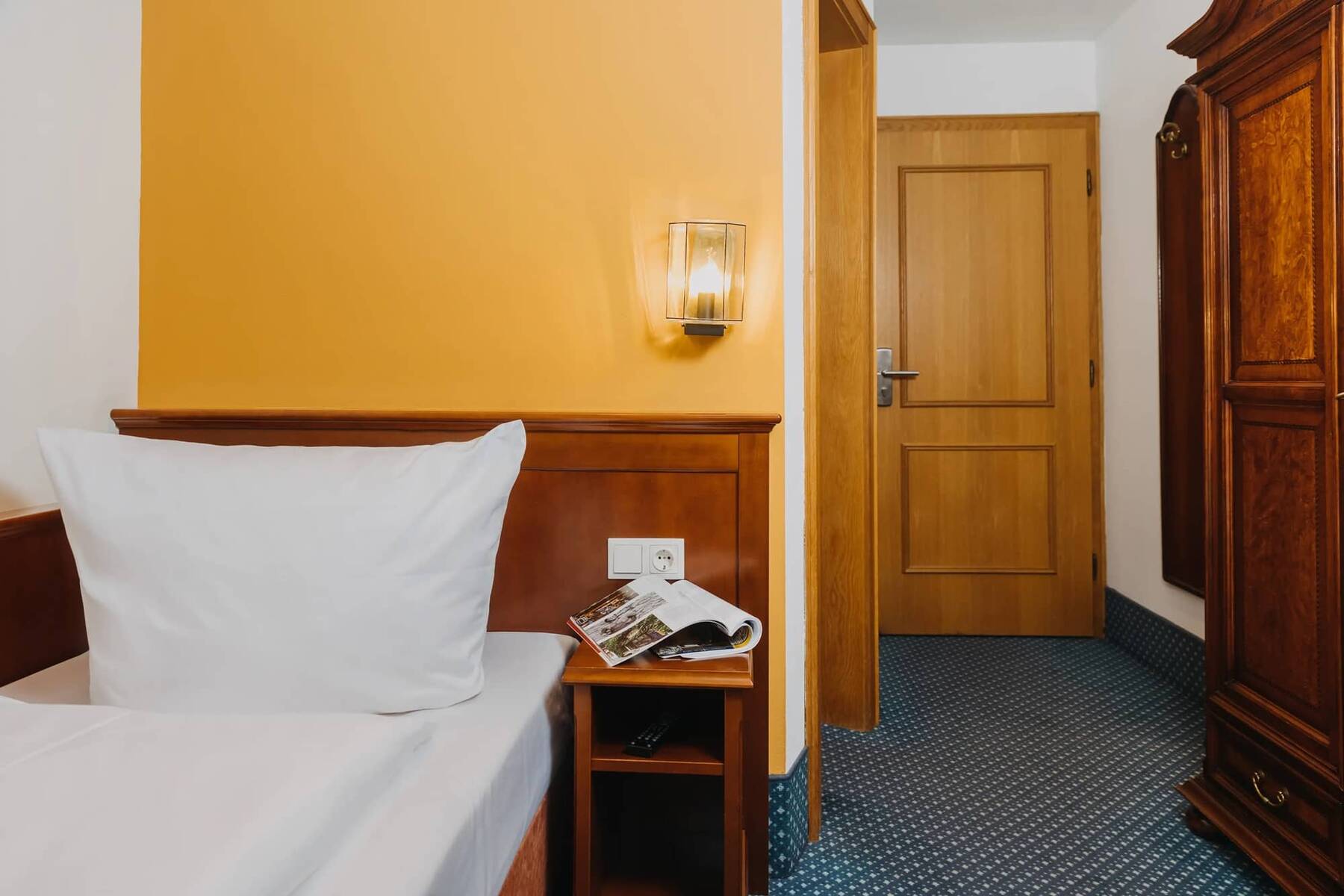 Einblick in ein gemütliches Einzelzimmer im Hotel, ausgestattet mit einem großen Kissen, einem Nachttisch und Holzmöbeln.