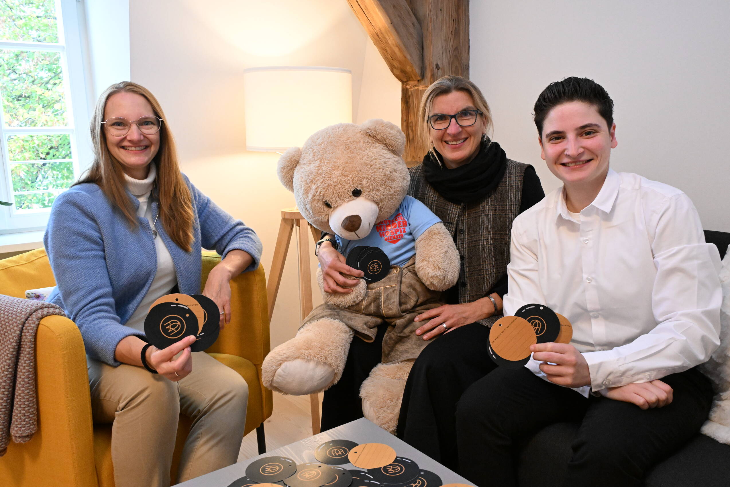 Drei freundliche Personen im Familienhotel halten Karten und sitzen mit einem großen Teddybären auf einem Sofa.